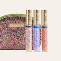 Three LipSense gloss tubes with ornate gold caps in front of a multicolored glitter clutch