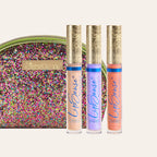 Three LipSense gloss tubes with ornate gold caps in front of a multicolored glitter clutch